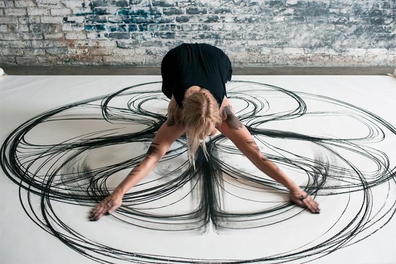 A photo of artist Heather Hansen on a white floor, using the movement of her arms to trace charcoal patterns on the ground.
