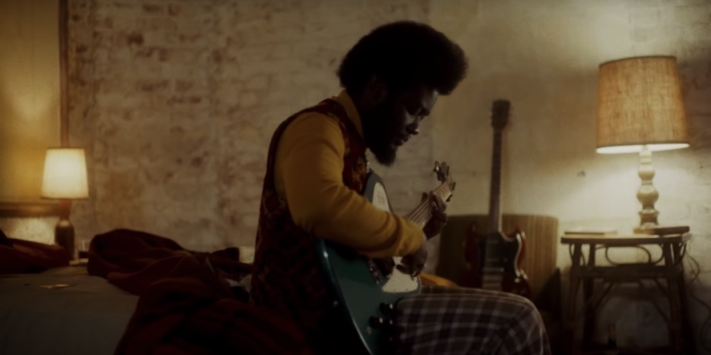 A photo still of Michael Kiwanuka sitting on a bed playing a guitar in the music video for Hero.