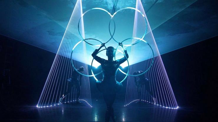 A photo still of three dancers from Quixotic. Two are standing underneath a row of laser lights, while the third stands in the centre holding five illuminated hula hoops.