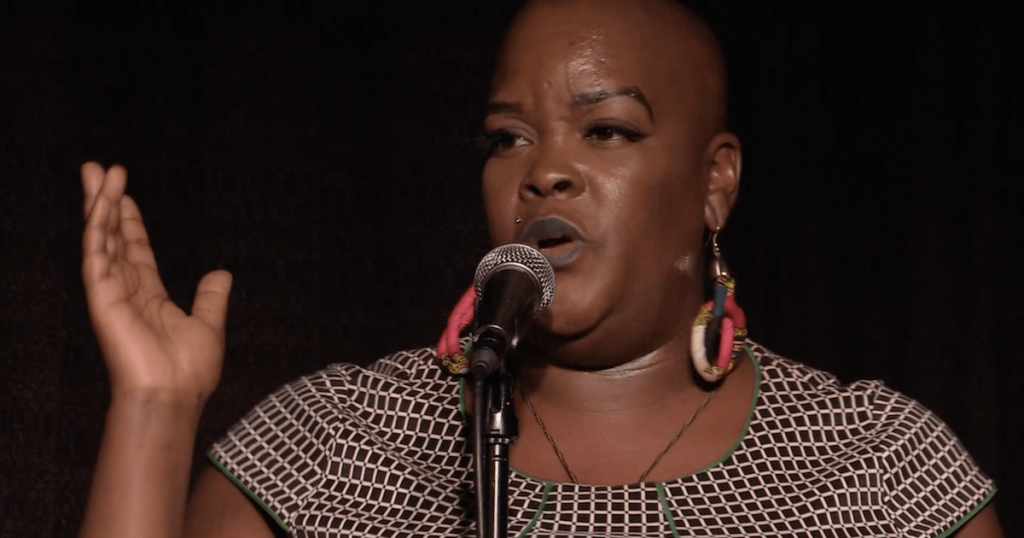 A photo of Sonya Renee Taylor speaking into the mic while performing The Body is Not An Apology.