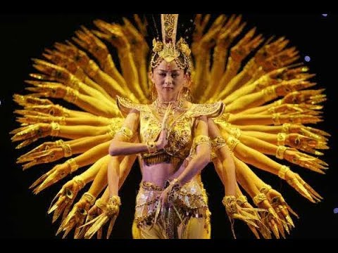 A photo still from a performance of the Thousand-Hand Bodhisattva. The dancer in front is looking straight at the camera, while dozens of arms appear at her sides from the dancers behind her.