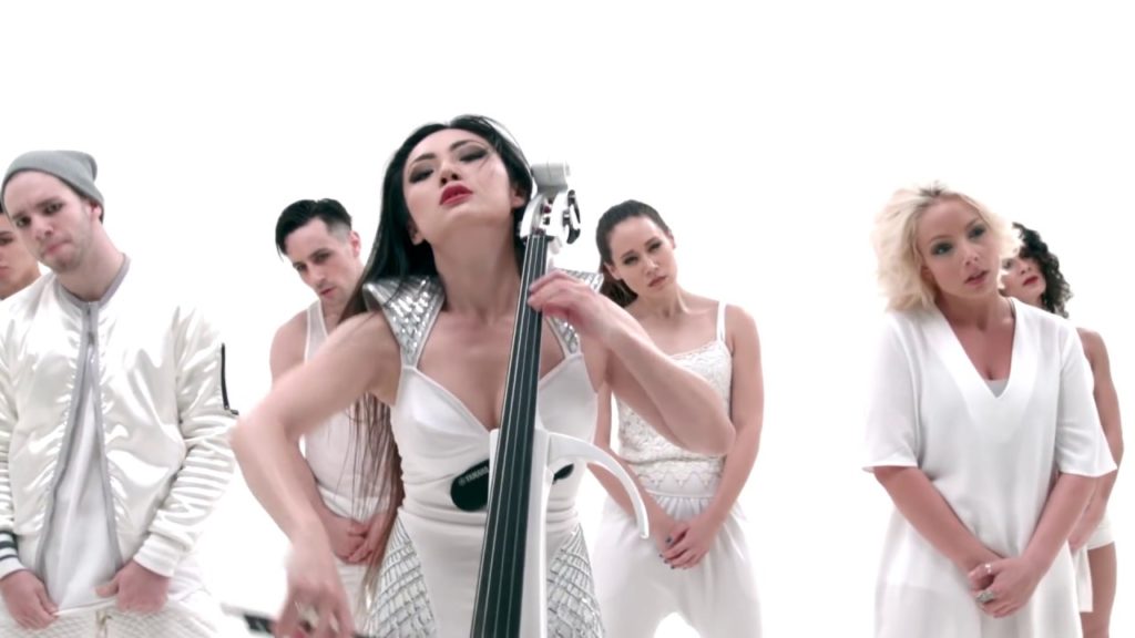 A photo still of Tina Guo from the music video of Crystallize. She is playing her cello with dancers behind her.