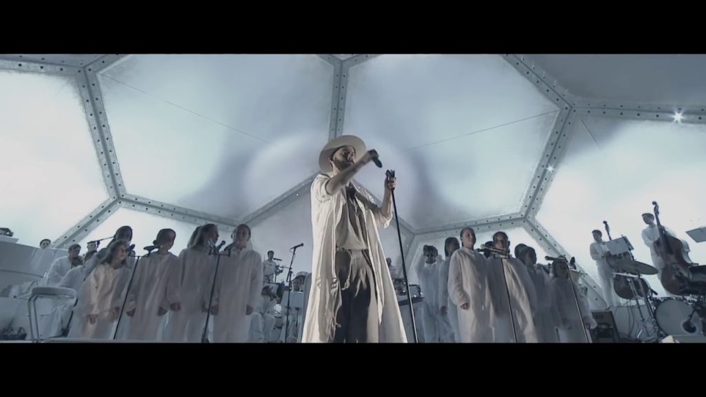 A photo of musician Woodkid centre stage during his live performance of Run Boy Run. He is surrounded by an orchestra and choir.