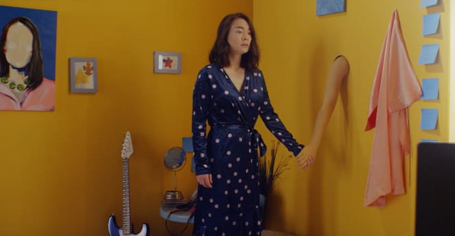 A photo still from the video for Nobody with singer Mitski holding the hand of an arm that is sticking out of a wall.