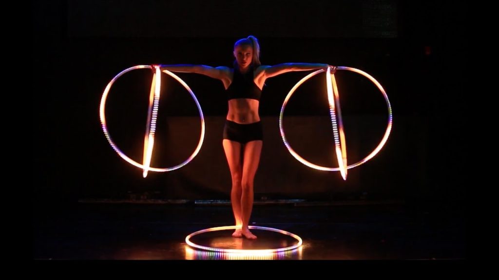 A photo of dancer Satya Bella looking straight into the camera, holding two LED hula hoops in each hand, and another at her feet on the floor.