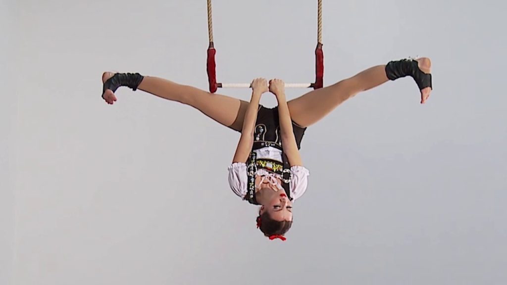 A photo of performer Ali Walker, mid-performance, hanging upside down from the static trapeze.