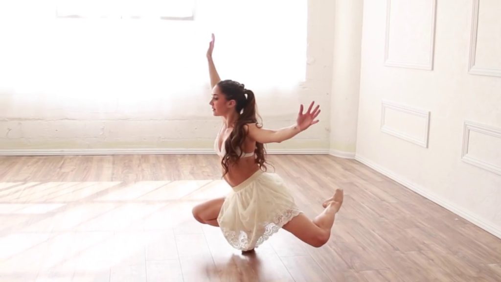 A photo still of dancer Audrey Case, mid-performance, squatting down on one foot with the other balanced behind her.