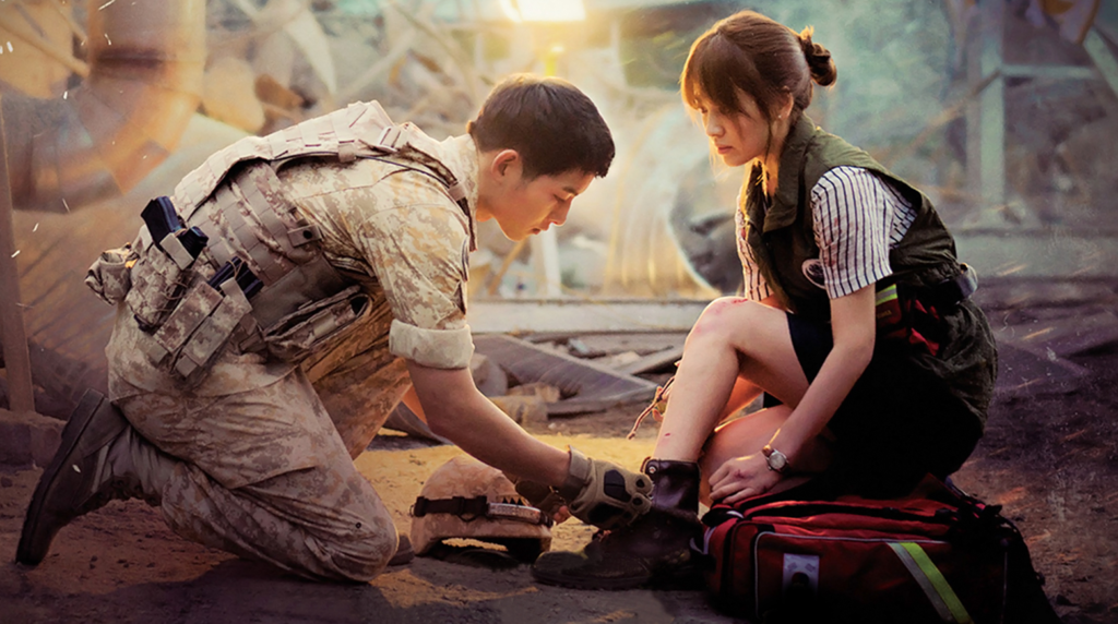 A promotional still from the TV series Descendants of the Sun, wherein lead actor Song Joong-Ki is tying the shoe of lead actress Song Hye-Kyo in the middle of a disaster zone.