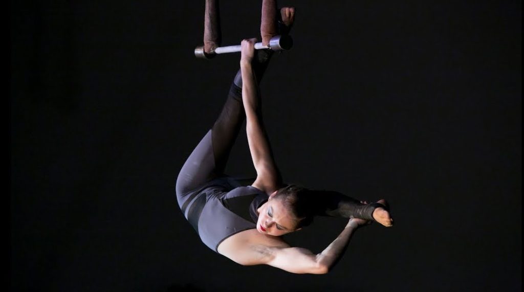 A photo still of acrobat Kae Henning, mid-performance, hanging by one arm from the static trapeze with one leg extended back behind her head.
