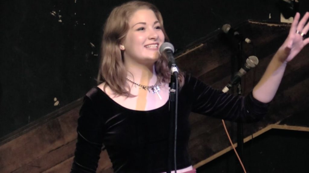 A photo of poet Blythe Baird, onstage, standing at a mic, mid-performance of When the Fat Girl gets Skinny