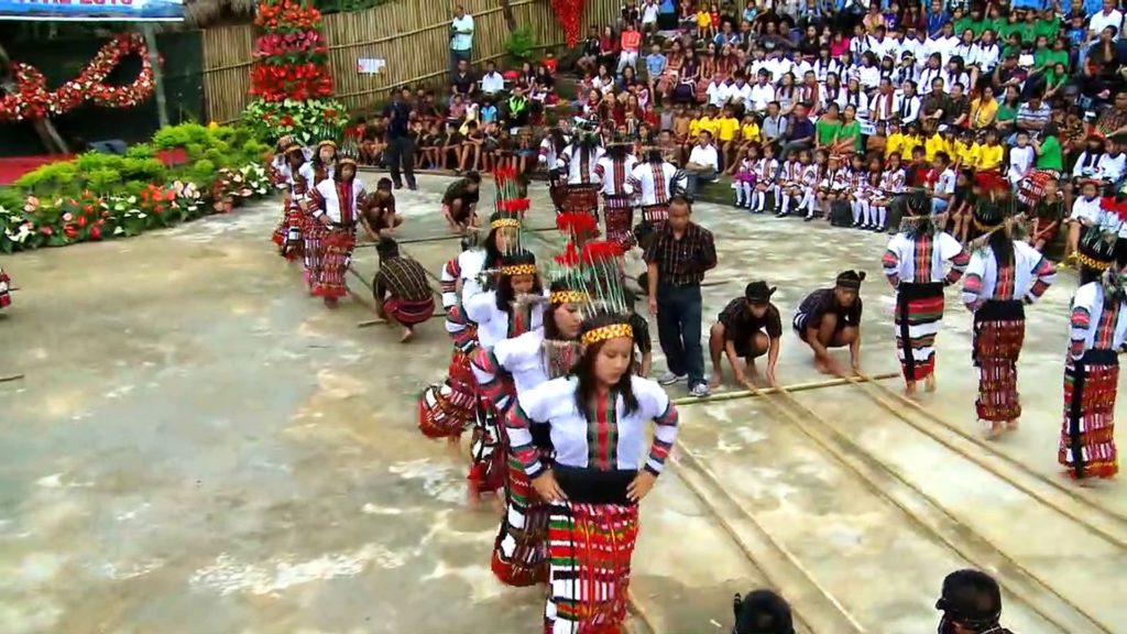 A photo still from a performance of the Cheraw dance, showing the female dancers standing in a line and the male performers holding the bamboo poles on the ground.