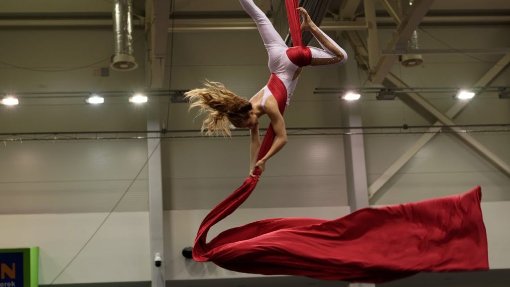 A photo of performer Krisztina Vellai, mid-performance, upside down in red silks.