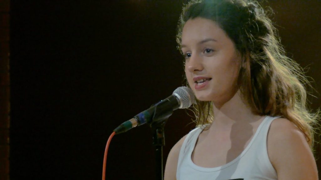 A close up photo of Olivia Gatwood standing at a microphone on stage.