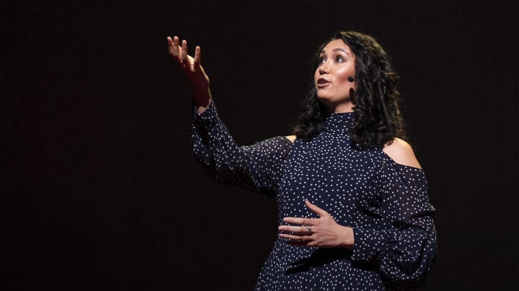 A photo of spoken word poet Sarah Kay, onstage, mid-performance, with one hand raised in the air.