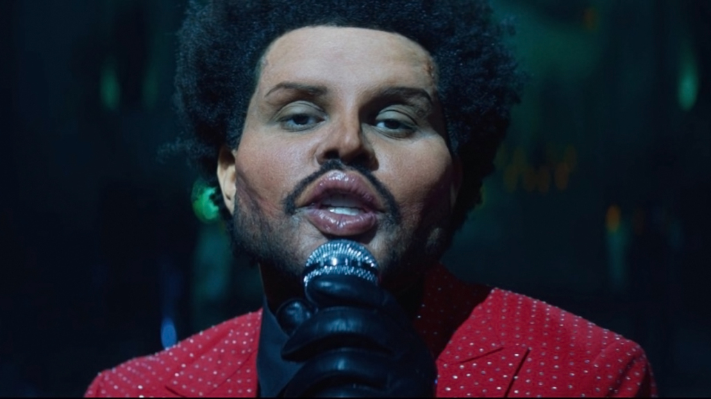 A close up photo of The Weekend from the music for Save Your Tears. He is holding a mic and looking straight into the camera.