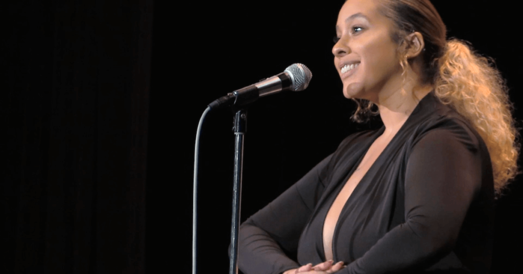 A photo of poet Anita D, onstage in front of a mic, mid-performance, holding her hands together in front of her.