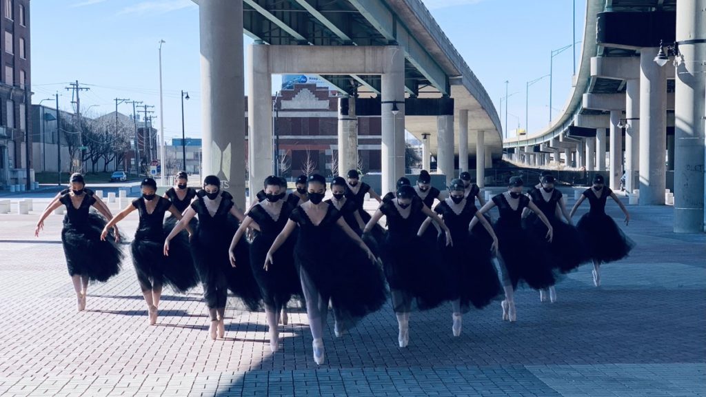 A photo of the dancers of Crescendo Conservatory, dancing under an overpass in an urban setting. They are all wearing classical black ballet dresses and black face masks.