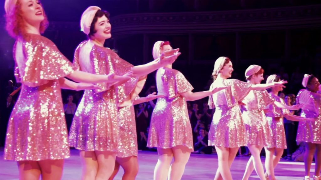 A photo of the dancers from JazzMAD, onstage, in a line, holding out their hands to the audience, wearing pink sequinned dresses.