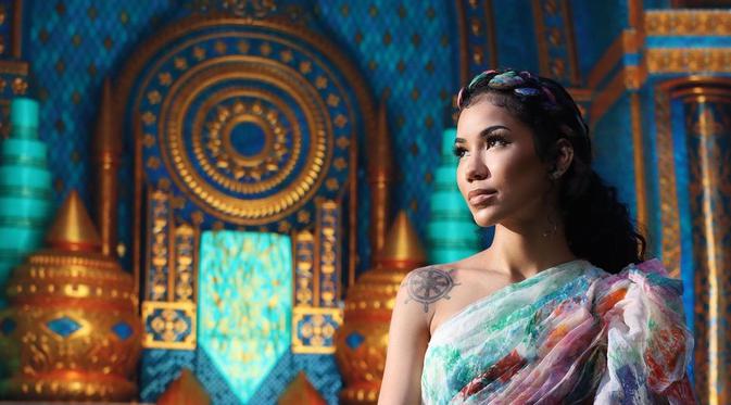 A photo still of singer Jhené Aiko from the music video for Lead the Way. She is standing in a colourful room, wearing an off the shoulder multi-colour dress and headband, looking off to the left.