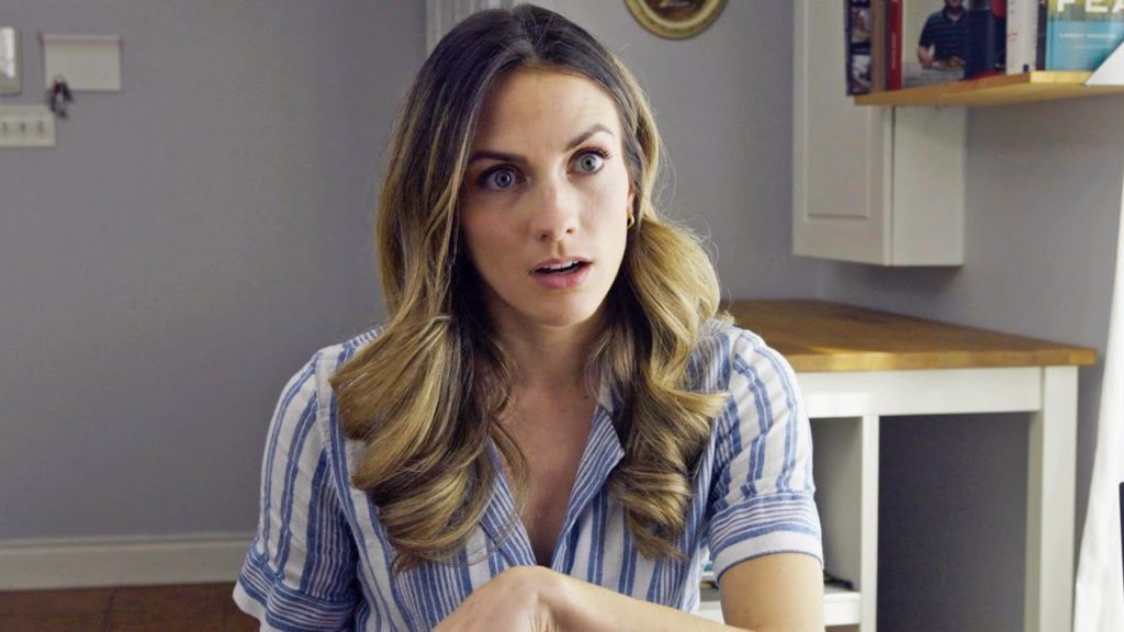 A photo still of comedian Julie Nolke sitting at a kitchen table looking quite perplexed.