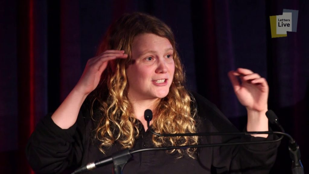 A photo of poet Kae Tempest, onstage in a black dress, standing in front of a lectern with two microphones, hands in the air and mid-performance of Brand New Ancients.