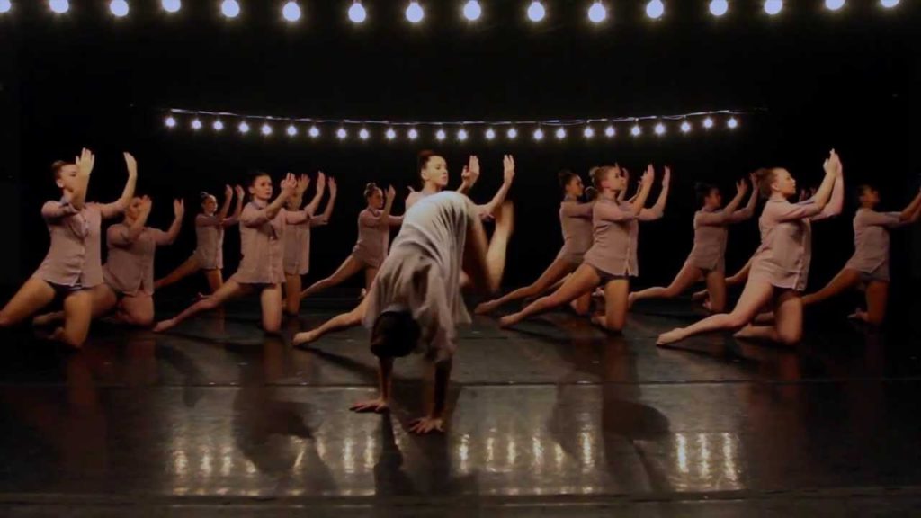 A photo still of Kate Jablonski and the company of Beyond Words mid-performance of their piece "In This Shirt". About a dozen dances in beige costumes are on their knees onstage with their hands raised in the air.