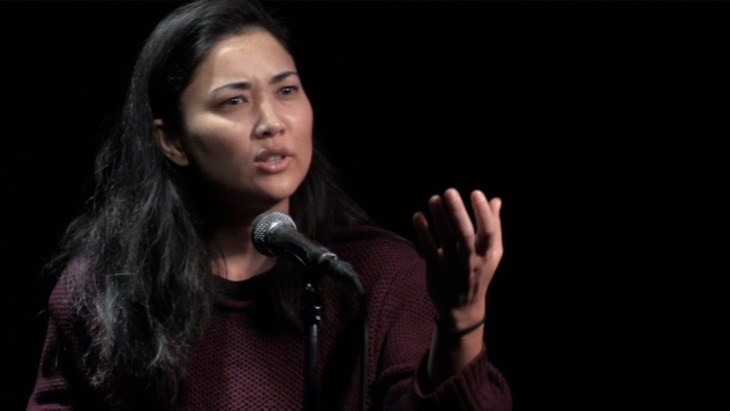 A photo of poet Leah Anderson onstage, mid-performance, standing in front of a mic with one hand raised.