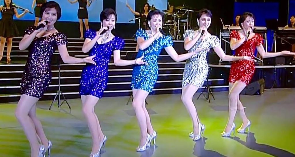 A photo of the five singers of the Moranbong Band, all wearing brightly coloured sequinned dresses, mid-performance, all holding microphones, standing in a line onstage.