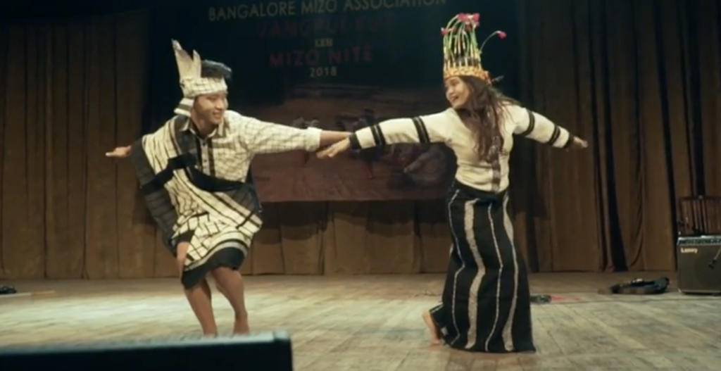 A photo of dancers Hmangaihsanga & Nancy onstage, standing side by side, mid-performance, with their hands extended to their sides, looking at each other.