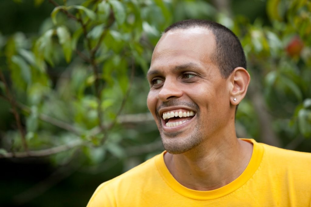 A close up photo of poet Ross Gay, wearing a bright yellow t-shirt, looking off to the side, and smiling a wide smile.