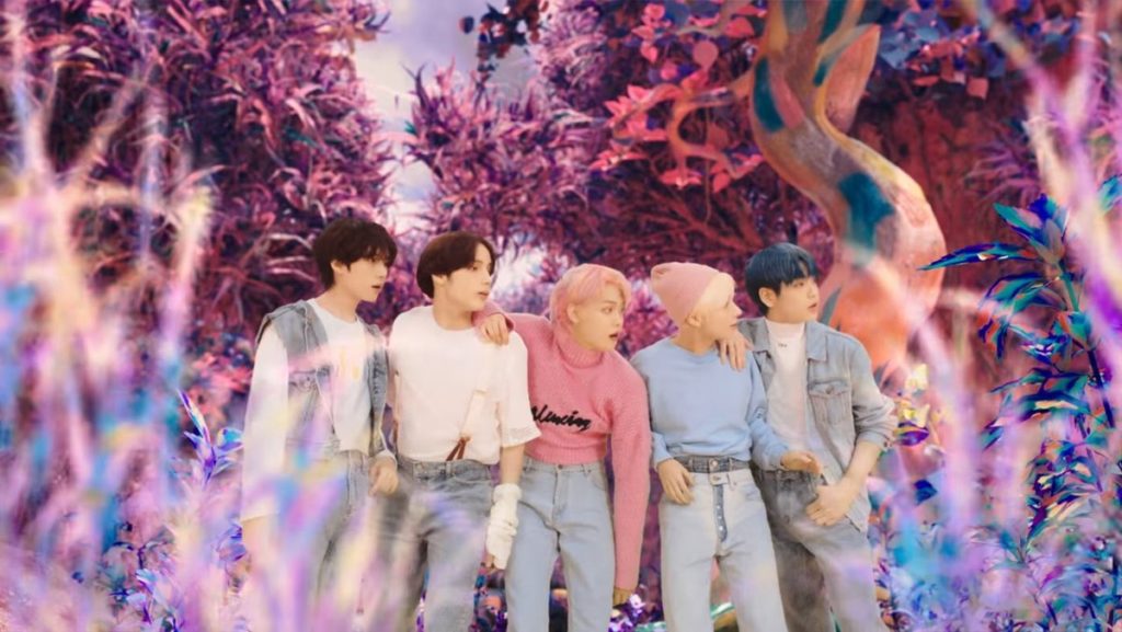 A photo still of the five members of TXT from the music video of Blue Hour. They are all looking off to the right and are surrounded by a colourful fantasy forest.