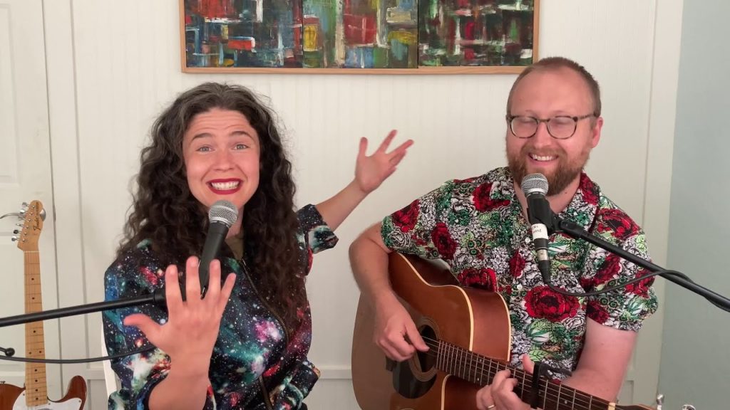 A photo of the Bengsons, singing directly into the camera with two mics and a guitar in Shaun's hands in a nondescript white walled room.