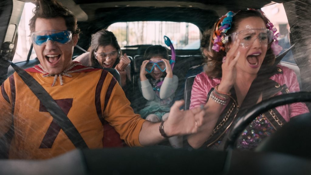 A photo still from the movie Yes Day, showing a family in a car, all dressed in outrageous outfits, going through a carwash with the windows down and getting soaking wet.
