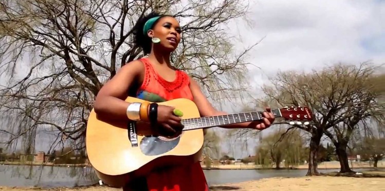 A photo of singer Zahara standing in a park, holding a guitar. There is a lake and trees in the background, and she is wearing a red dress and a turquoise headband.