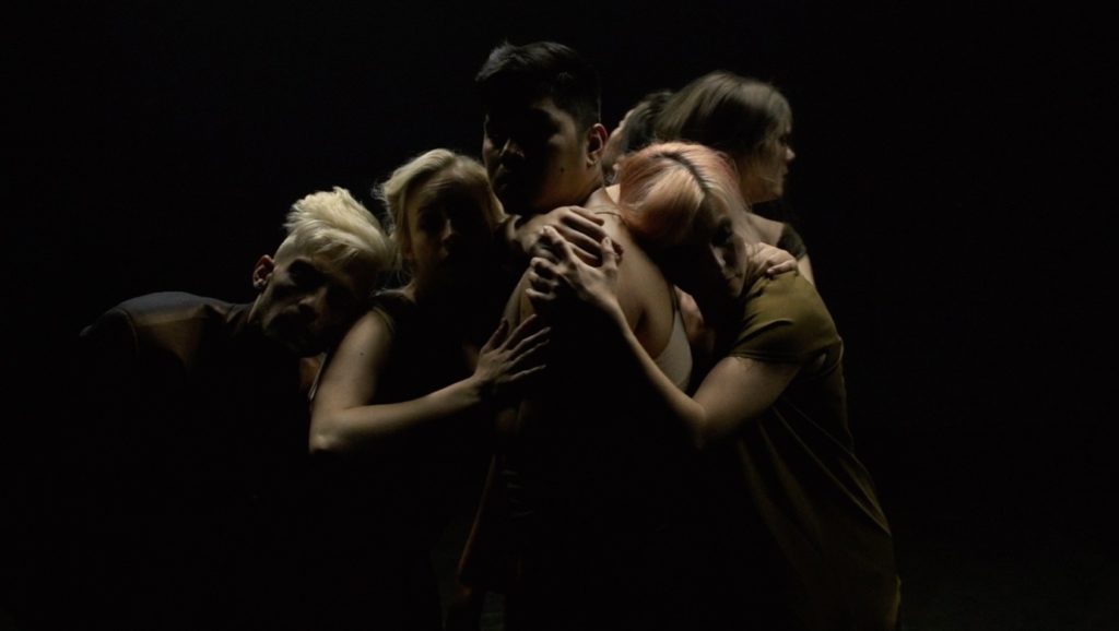A photo still from the film BLINK by the LA Contemporary Dance Company of several dancers, all huddled together, their faces shrouded in shadow.