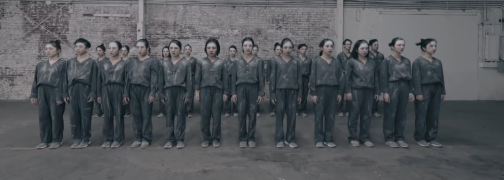 A photo still of the dancers of Chibi Unity, standing in two straight lines, one behind the other, all dressed in grey outfits with white chalk on their faces, facing the camera.