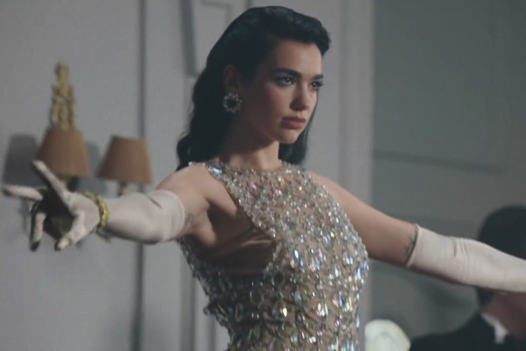 A photo still of singer Dua Lipa from the video for We're Good. She is styled from the turn of the century and posing onstage while she sings.