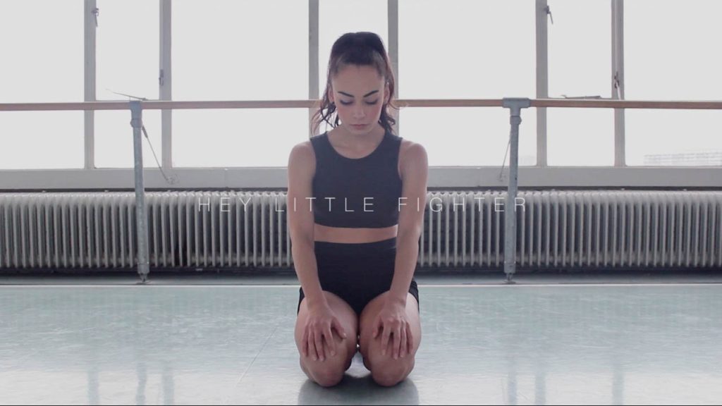 A photo still from the video Hey Little Fighter of dancer Raifa Bazzi, kneeling on the floor, hands palm down on her thighs, eyes closed and head tilted towards the floor.