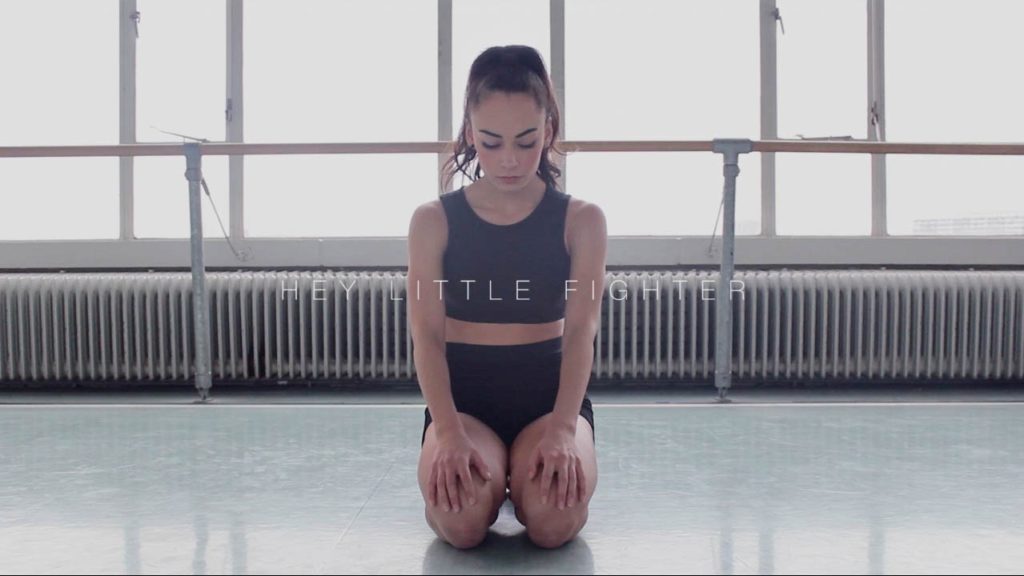 A photo still from the video Hey Little Fighter of dancer Raifa Bazzi, kneeling on the floor, hands palm down on her thighs, eyes closed and head tilted towards the floor.
