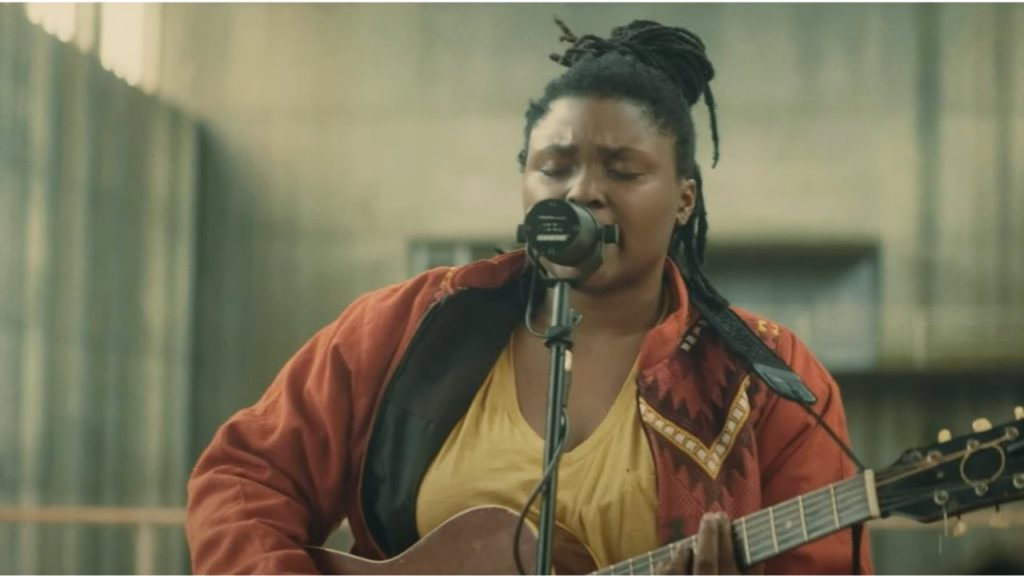 A photo of Joy Oladokun playing the guitar and singing into a mic with her eyes closed.