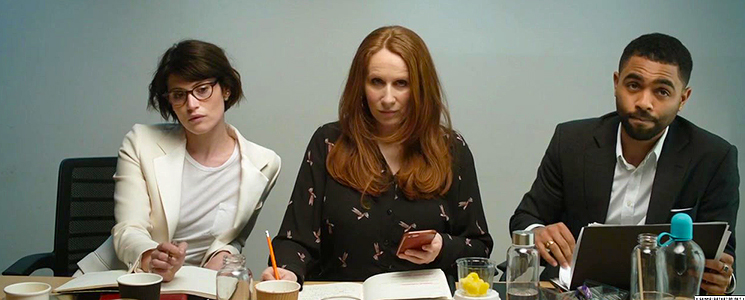 A photo of actors Gemma Arterton, Catherine Tate, and Anthony Welsh from the Leading Lady Parts video. They are sitting in a line, at a desk, facing the camera, with various notebooks, pens, and water bottles in front of them.