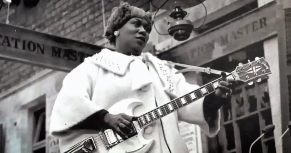 A black and white photo of Sister Rosetta Tharpe wearing a fancy white coat and holding an electric guitar.