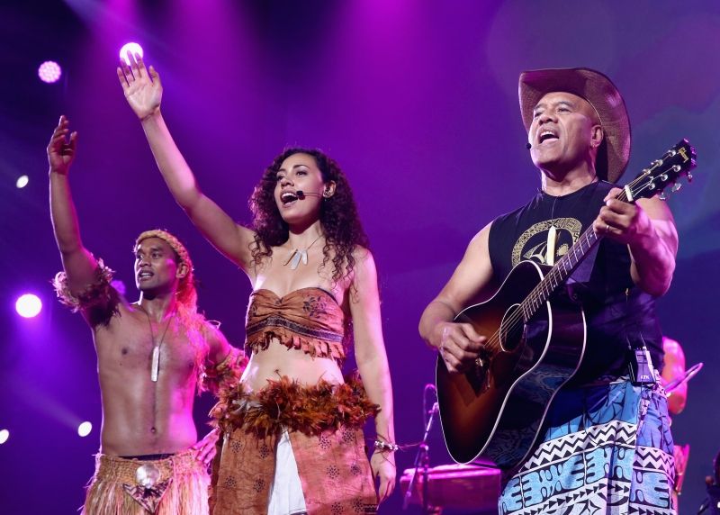 A photo of three members of Te Vaka, onstage, mid-performance, singing into mics.