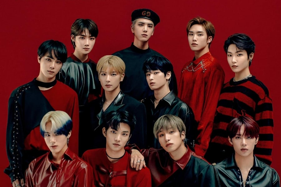 A promotional photo of the 11 members of The Boyz, all standing in front of a red backdrop, staring straight into the camera.