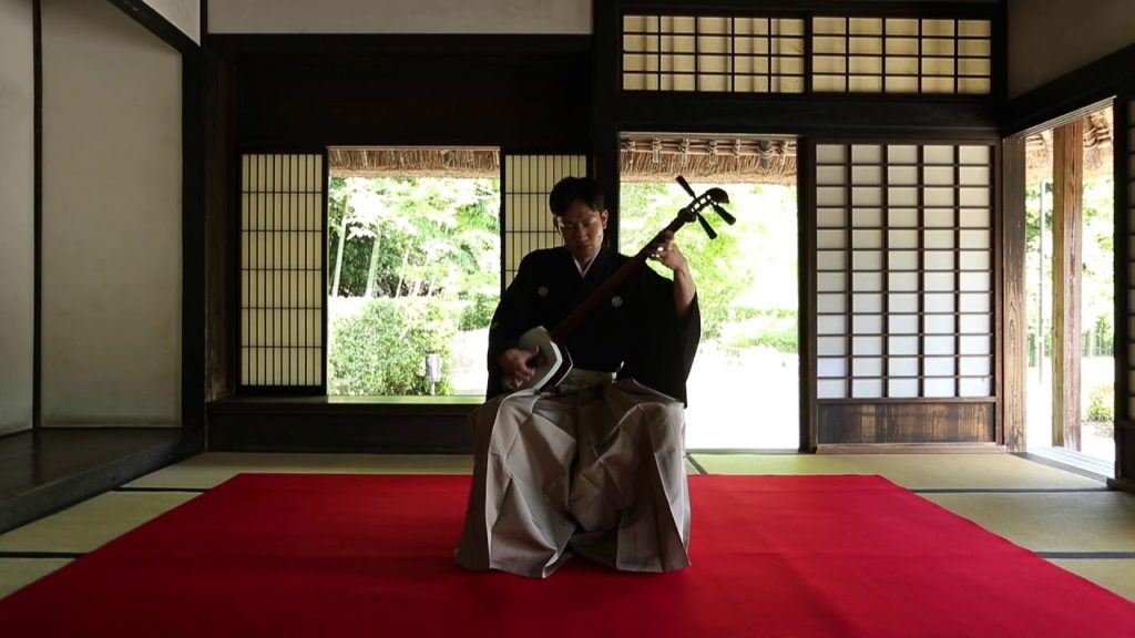 A photo of Tsugaru-shamisen player Masamitsu Takasaki sitting in a room of a traditional Japanese house with playing the shamisen.