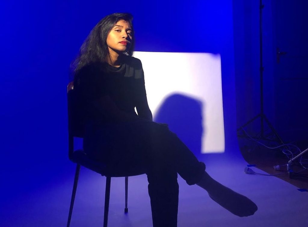 A promotional photo of Usha Jay, sitting on a chair, one leg crossed over the other, looking directly into the camera.