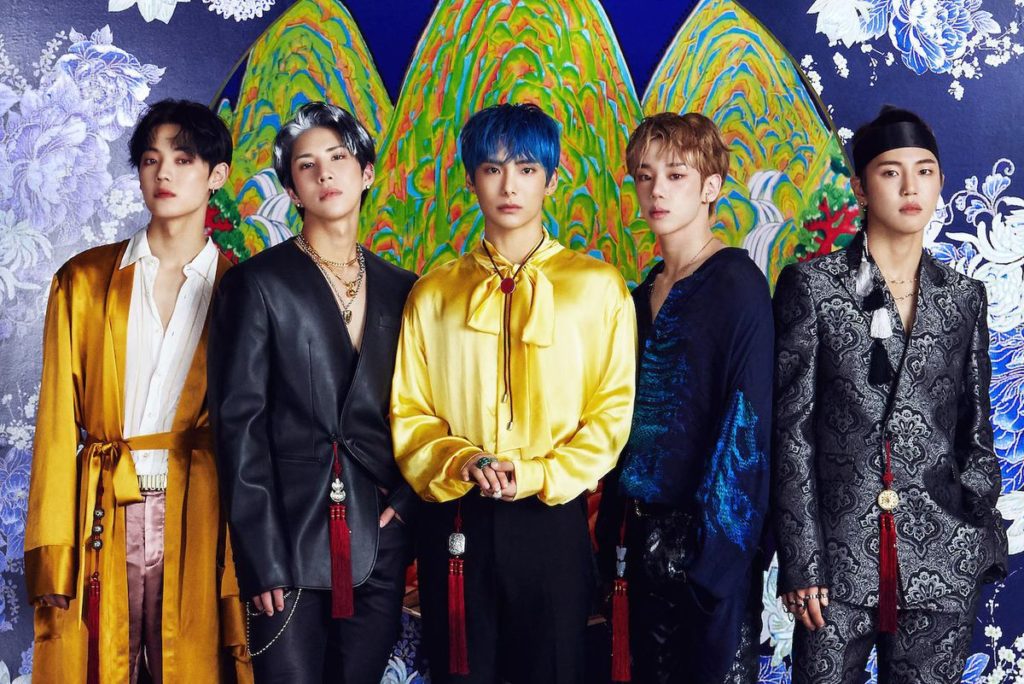 A promotional photo of A.C.E. All five members are standing in a line against a colourful wall and staring straight into the camera.