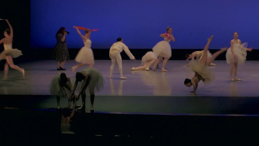 A photo still of Ballet Magnifique, mid-performance, with ballet dancers in traditional tutus in various states of disarray on a stage.