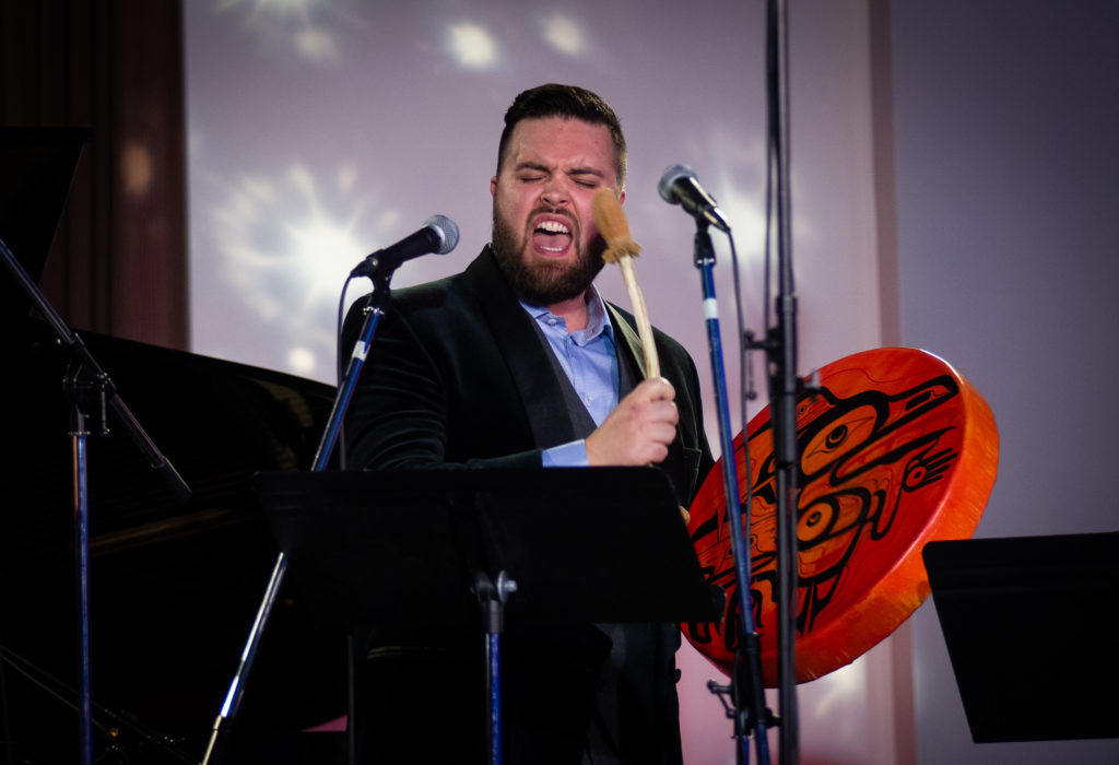 A photo of Corey Payette, on stage, mid-performance, singing into a mic and banging a drum that he is holding in his right hand.
