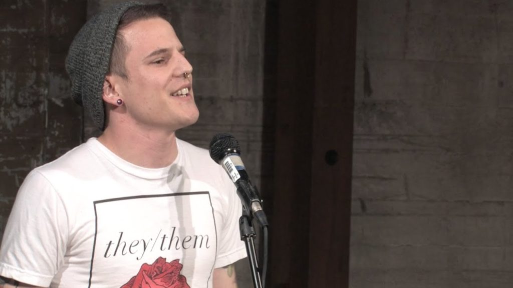 A photo of poet Jamie Mortara on stage, mid-performance, speaking into a mic, wearing a beanie and a white t-shirt with they/them pronouns.